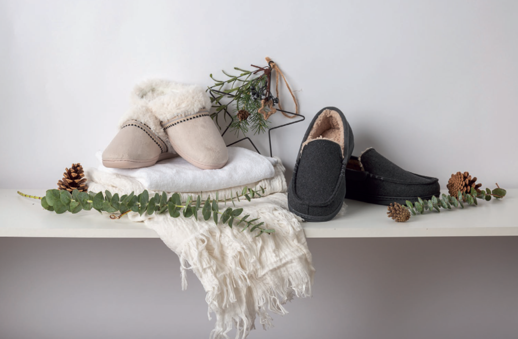 A pair of beige and grey slippers placed on a shelf, surrounded by pine cones and greenery, with a soft white blanket.